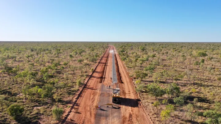 Drone view of a remote road construction site with heavy machinery compacting gravel on a partially sealed road