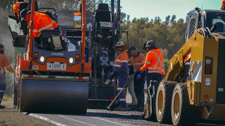 Road construction crew in high-visibility gear using heavy machinery, including a roller and skid steer, to lay and compact fresh asphalt