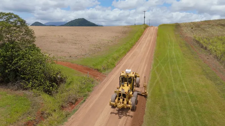 Yellow grader leveling a cassowary coast dirt road, surrounded by green grass and farmland under a partly cloudy sky