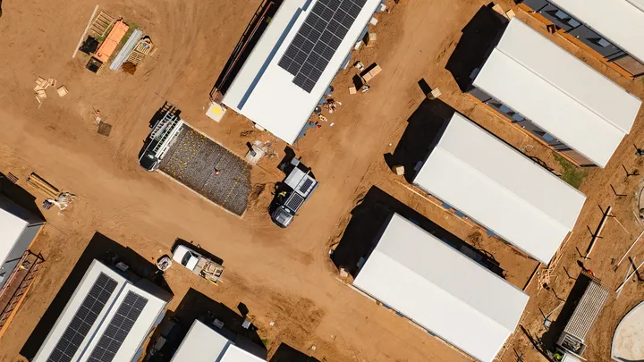 Overhead view of a construction site with multiple buildings arranged in rows, featuring white roofs, solar panels, and vehicles parked on dirt pathways.