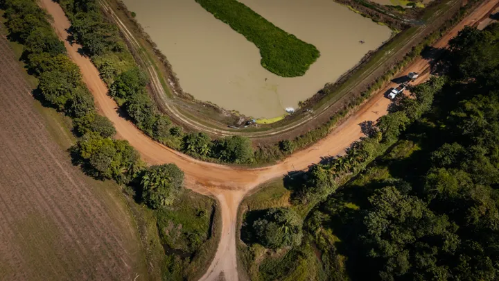 An aerial view of a regional cassowary coast dirt track intersection bordered by lush trees, with a paddock on one side and an aquaculture pond on the other.