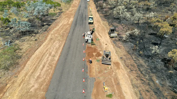 Drone view of a rural road construction site with workers, heavy machinery, and traffic cones, alongside a recently burned landscape.