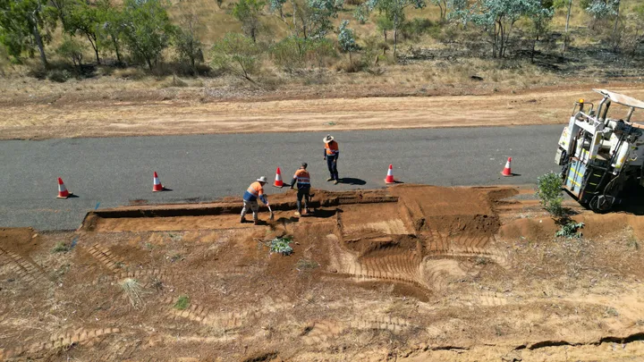 Road construction workers in high-visibility gear excavating a roadside trench with heavy machinery