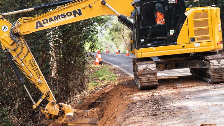 A yellow Adamson Earthmovers excavator digs along a roadside in a forested area, with traffic cones marking the work zone as part of a civil construction project.