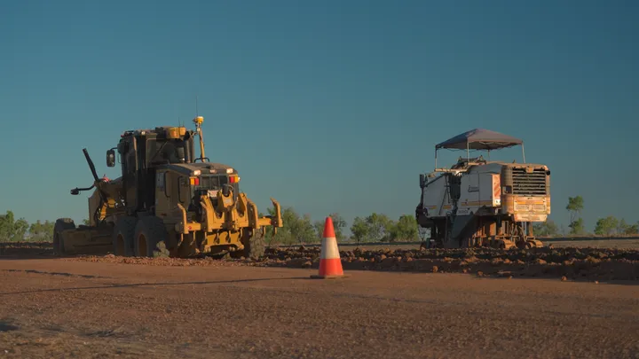 Grader and road stabilisation machine at work on a construction site, marked with a traffic cone for safety.
