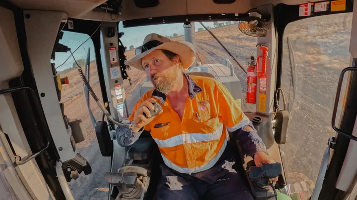 Construction worker operating grader inside a cab, communicating via a handheld radio on a road construction site