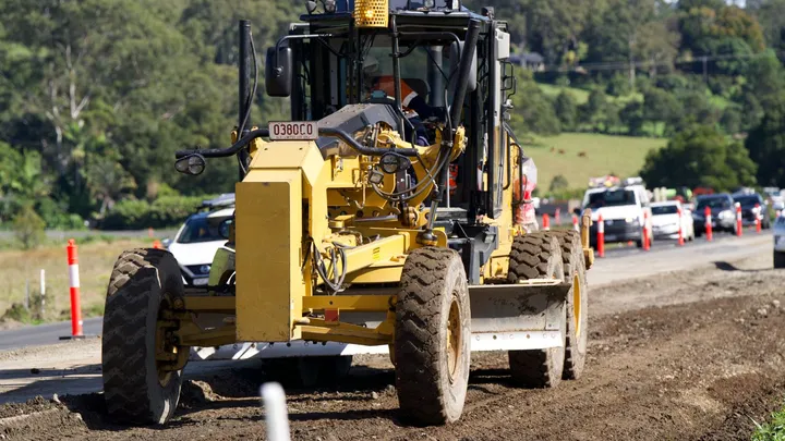 Road grader leveling the ground at a construction site, with traffic managed nearby using safety cones