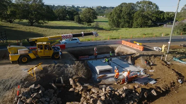 Construction workers and a mobile crane installing precast concrete culvert components at a roadwork site.