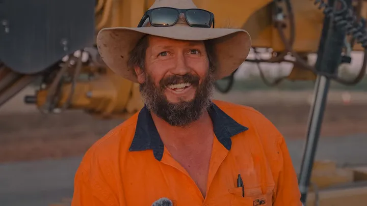 A cheerful Durack worker wearing a wide-brimmed hat and sunglasses on his head smiles while dressed in high-visibility orange gear, standing in front of civil construction machinery.