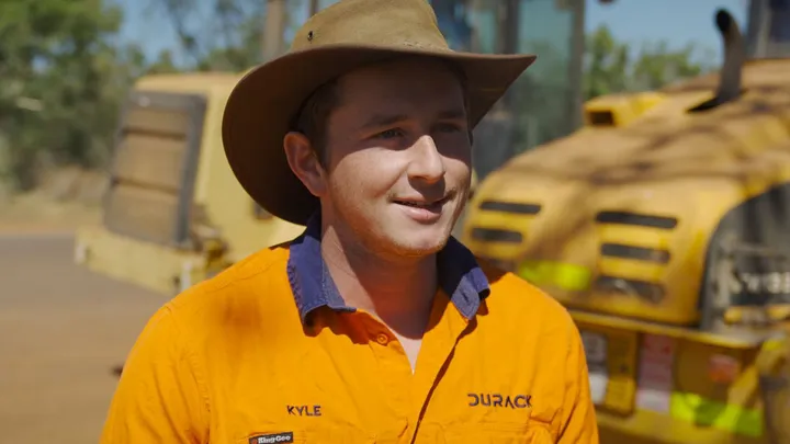 Kyle, wearing a wide-brimmed hat and a Durack high-visibility uniform, smiles confidently with construction equipment in the background.