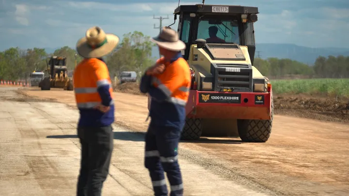 Two contractors in high-visibility uniforms discuss progress on a road construction site, with heavy machinery working in the background as part of a regional infrastructure project.